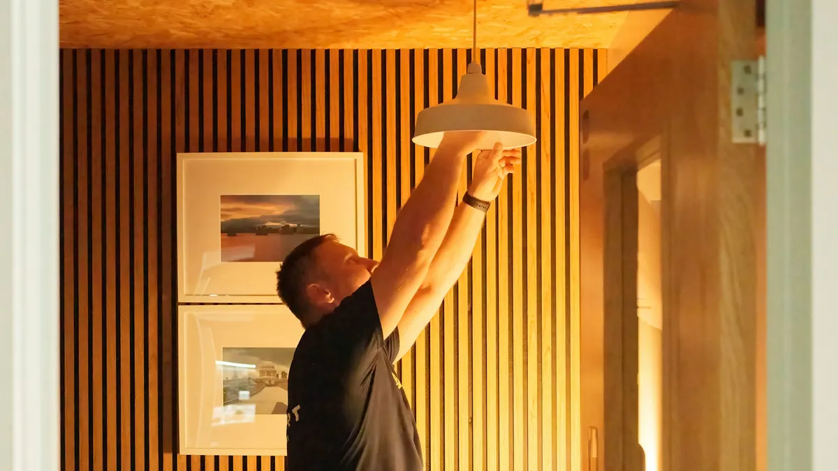 Vakman met gereedschapstas hangt een witte hanglamp op in een kamer met houten lamellenwand